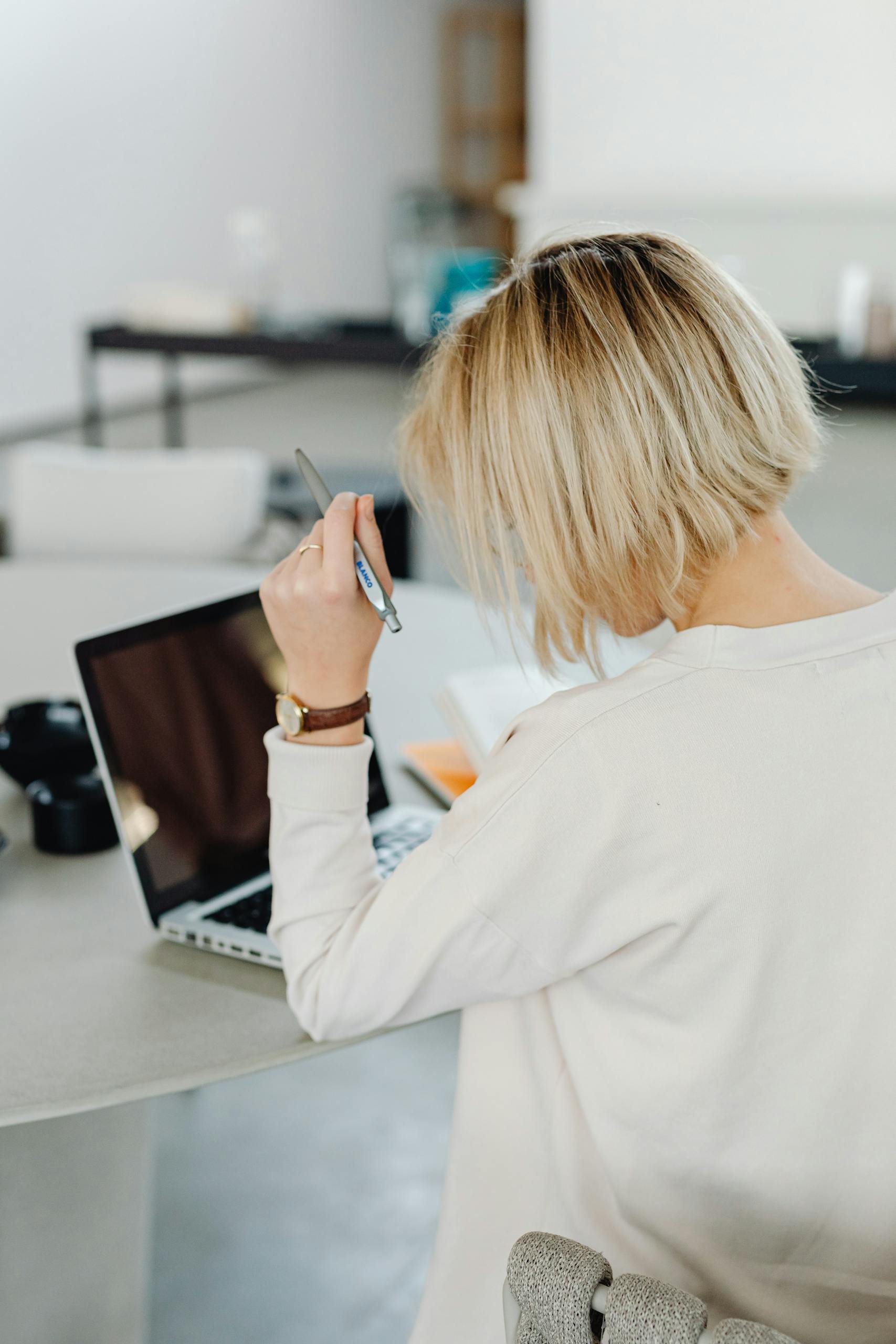 Blonde woman with short hair holding pen while working on a laptop at home, back view.